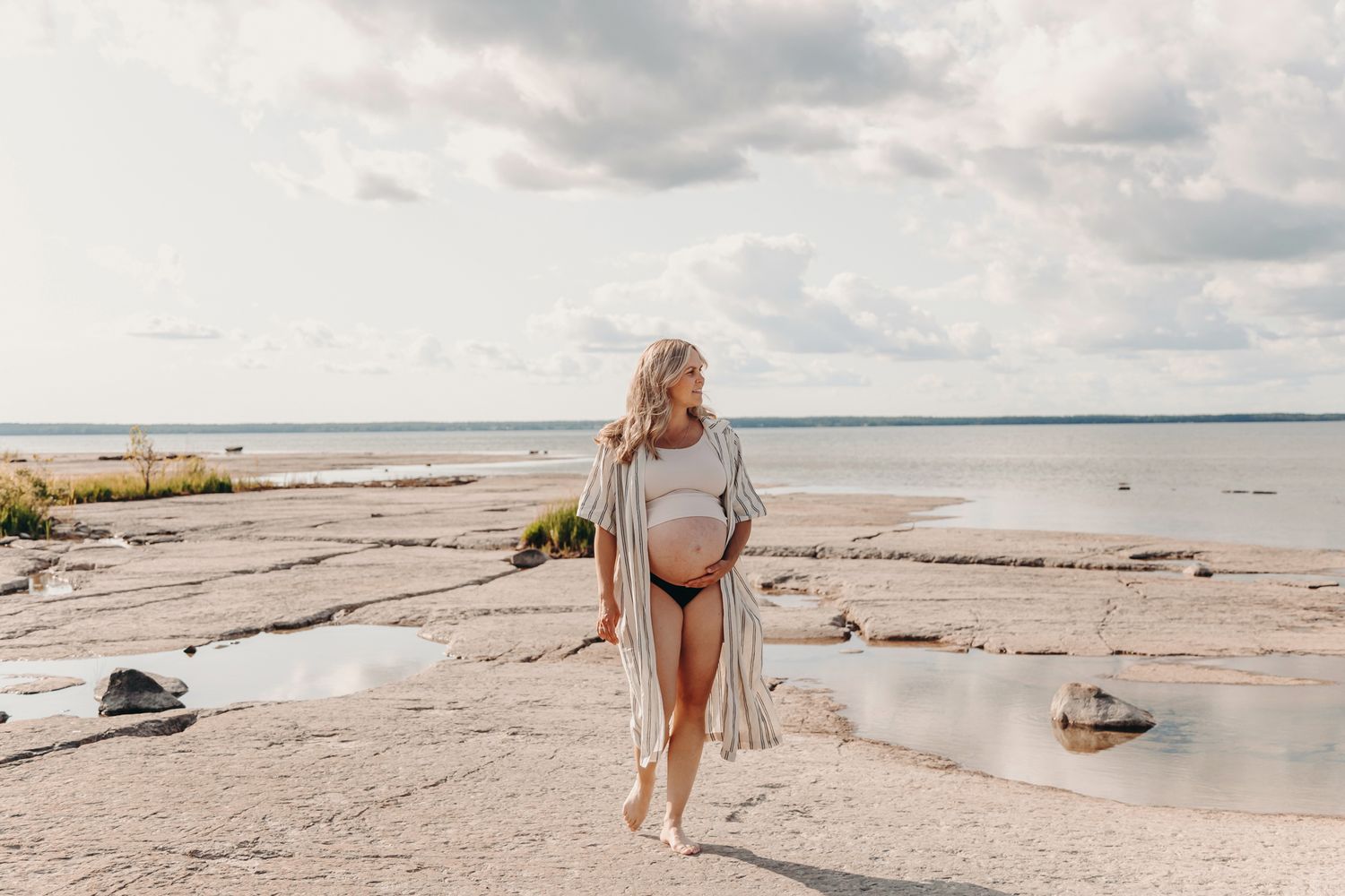Woman in white dress walks along scenic coastal beach with scattered driftwood and calm waters on a cloudy day.