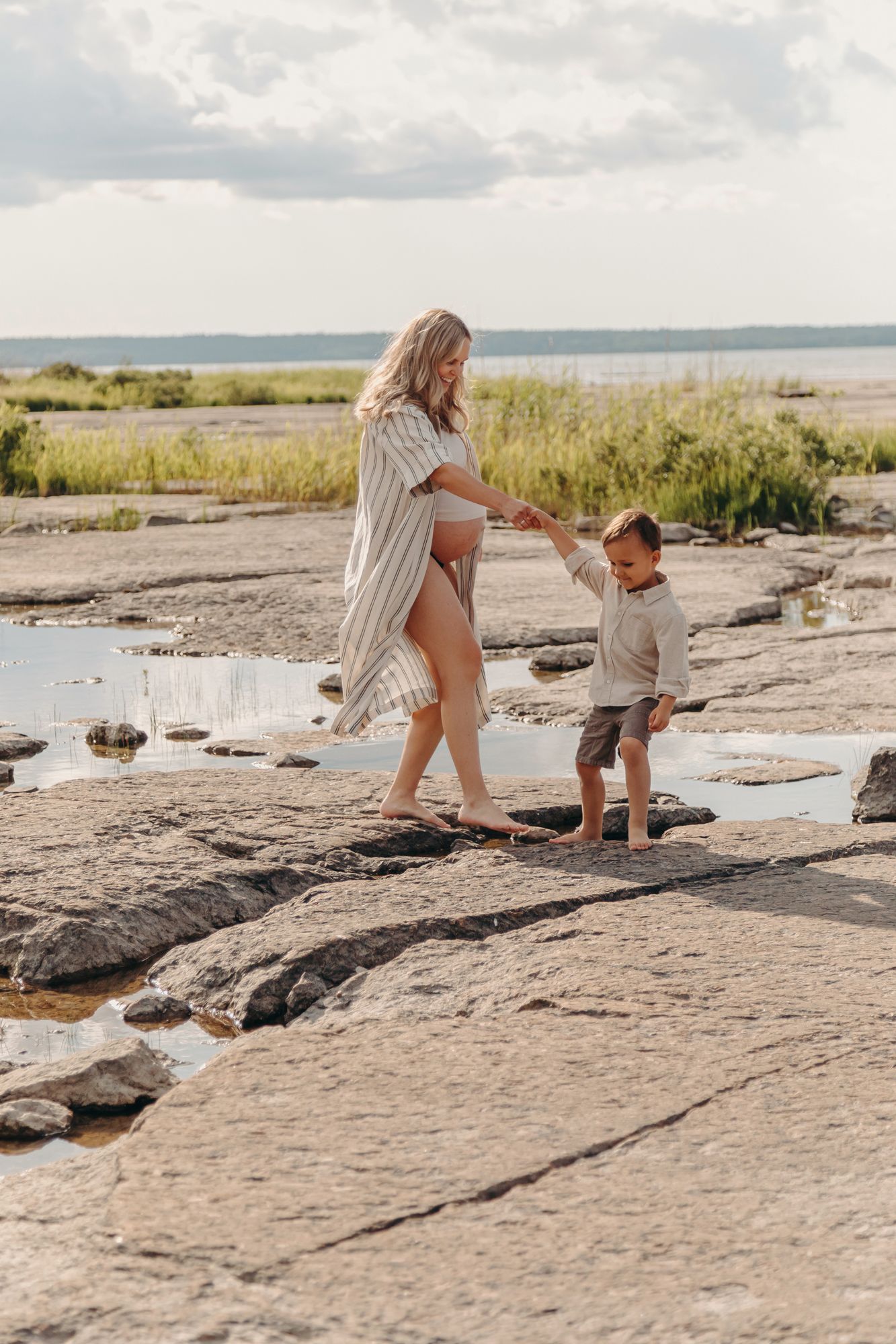 A parent and child explore rocky coastal terrain together on a sunny afternoon by the water's edge.