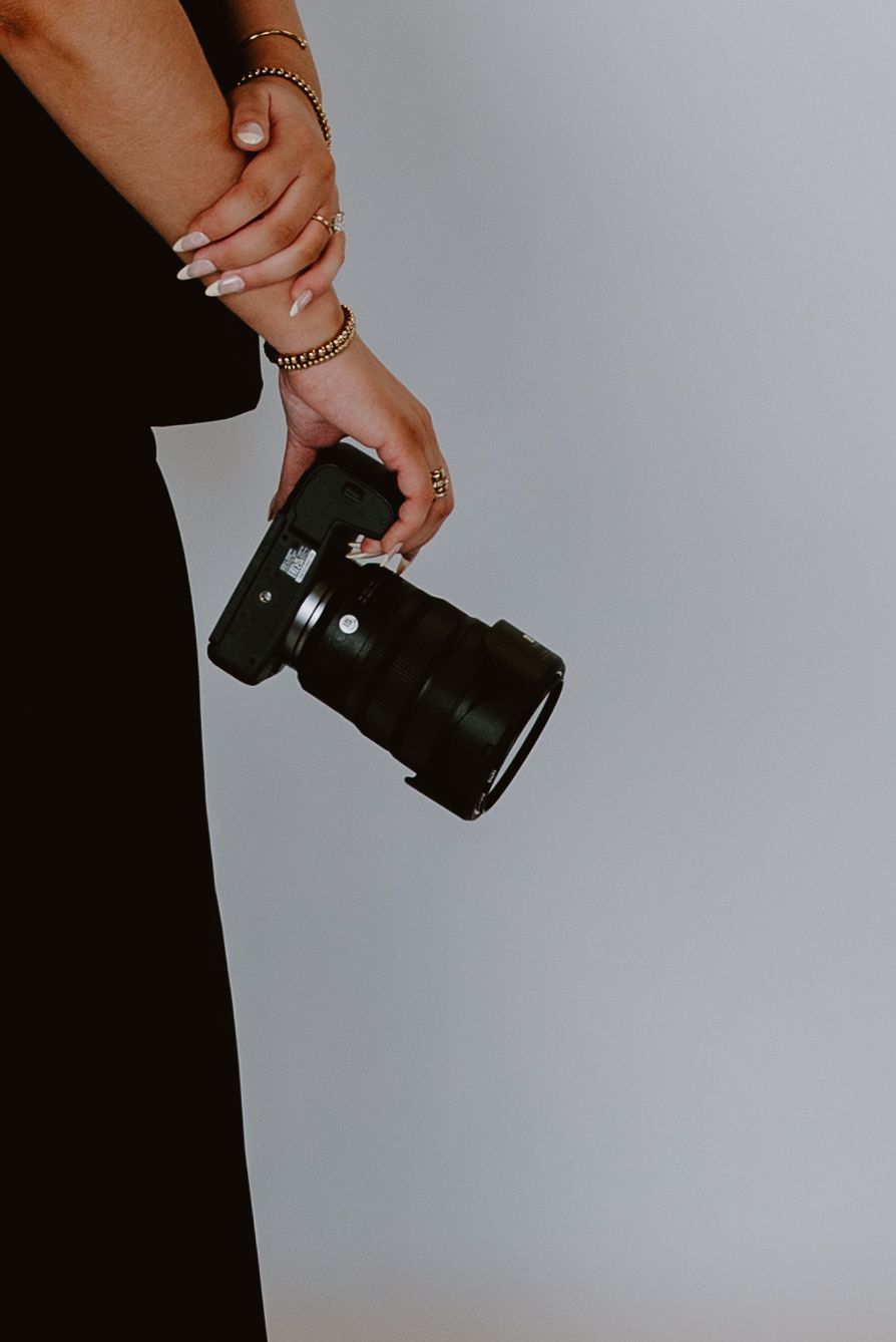 Close-up of woman in black dress holding professional DSLR camera with gold bracelets and rings.