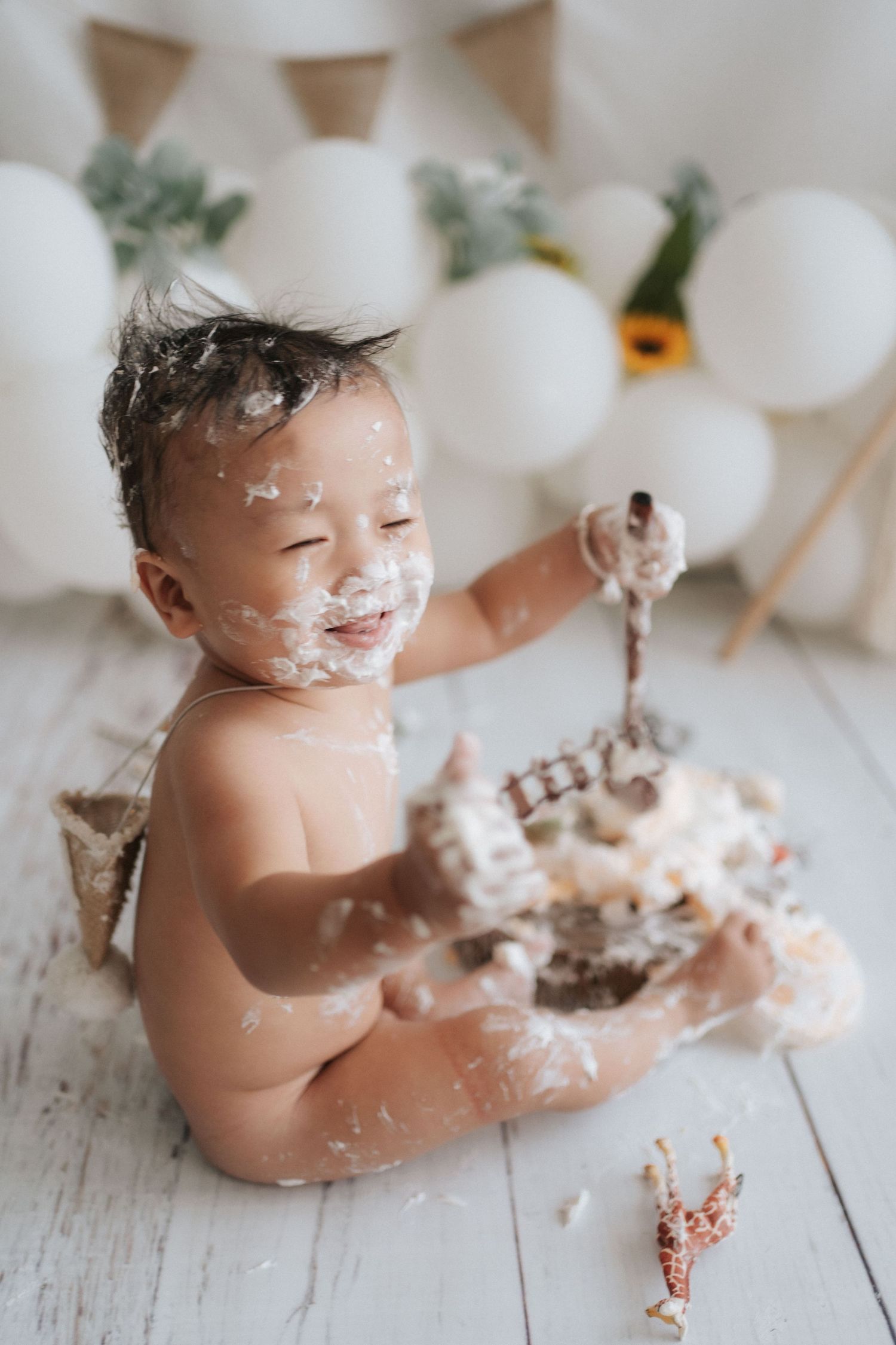Baby sitting on white wooden floor enjoying birthday cake smash photoshoot with white balloon decorations.