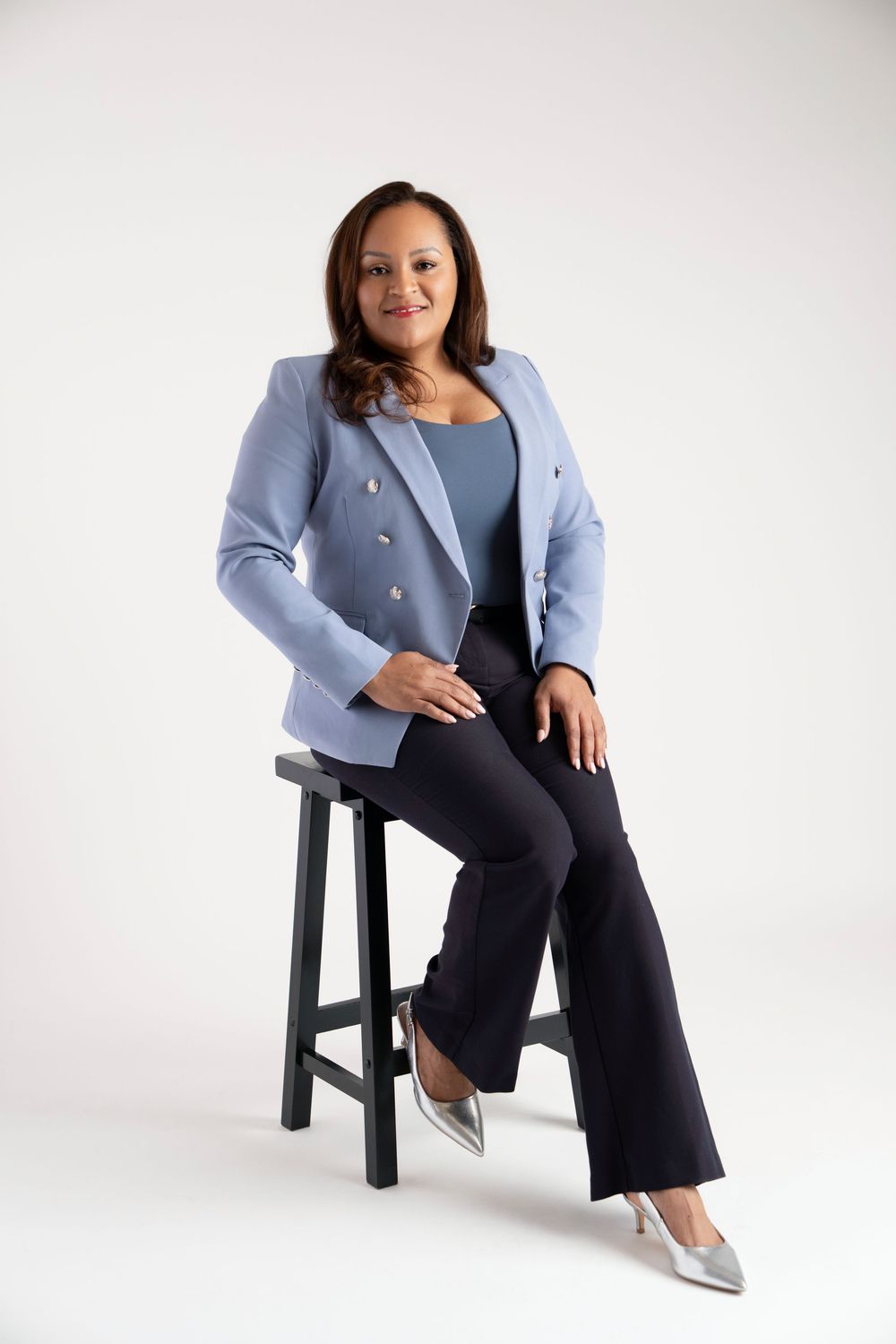 Professional wearing a light blue blazer and dark pants sits confidently on a black stool against white backdrop.