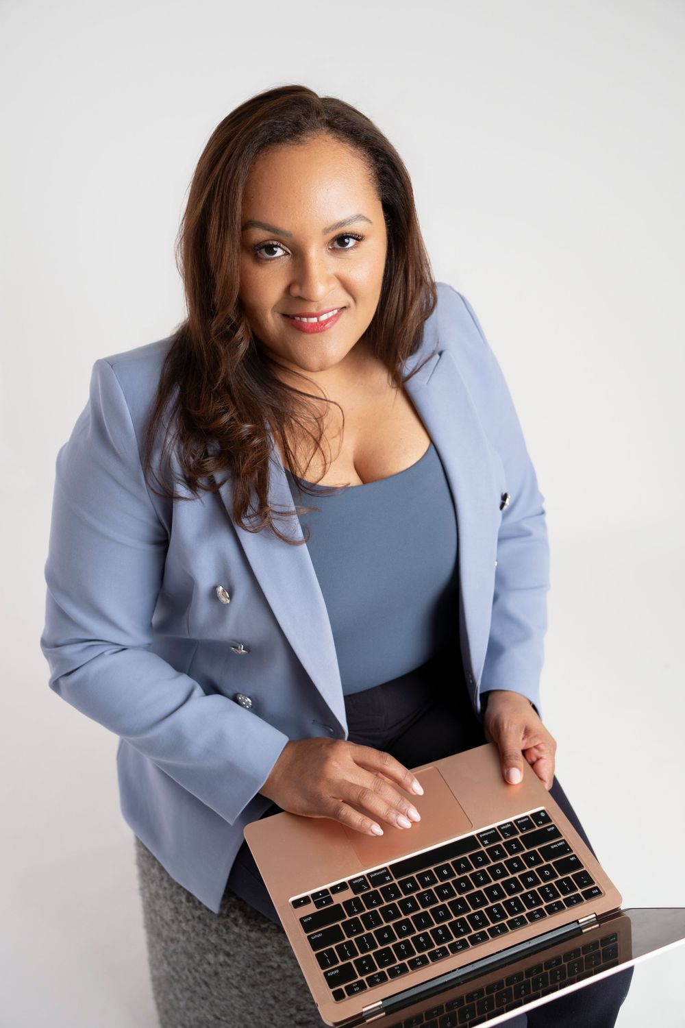 Professional wearing a light blue blazer smiles while working on a laptop against white background.