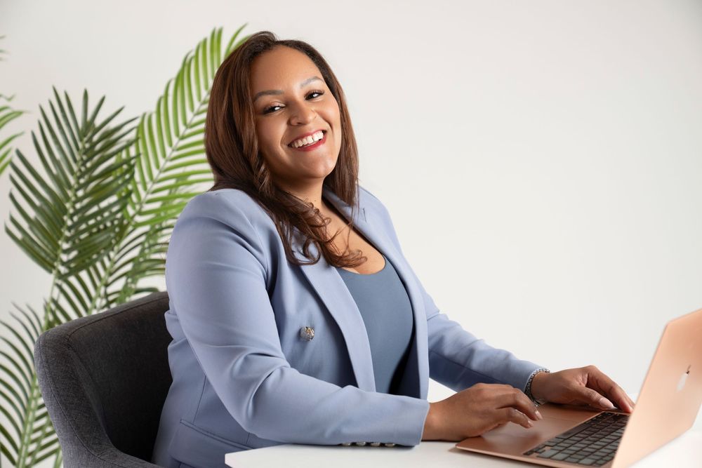 Professional in light blue blazer sits at desk with laptop and tropical plants in modern office setting.