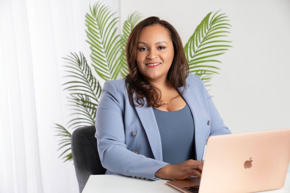 Professional wearing a light blue blazer works on laptop in bright modern office setting.