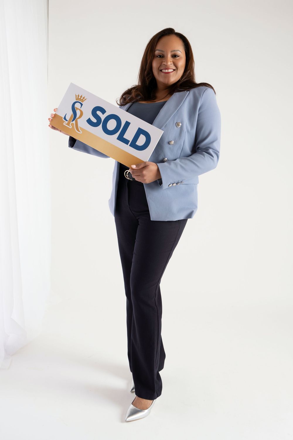 Real estate agent in light blue blazer and navy pants holds SOLD sign in professional photoshoot series.