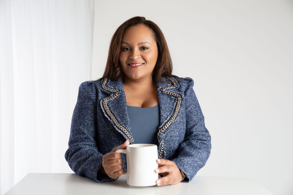 Professional in a navy tweed blazer holds a white coffee mug while sitting against a light colored background.