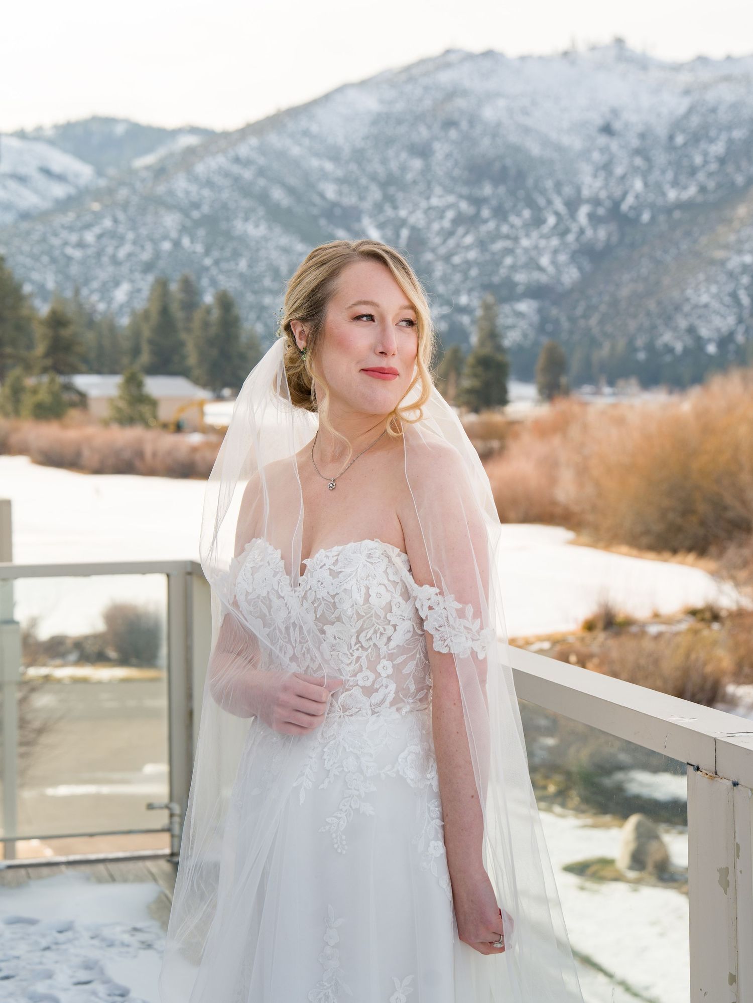 A bride in a white lace wedding dress poses against a snowy mountain backdrop on a winter day.