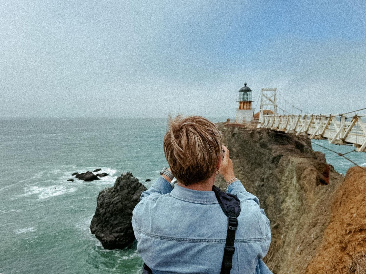 Person with short hair photographing a coastal lighthouse from rocky cliffs overlooking the ocean.