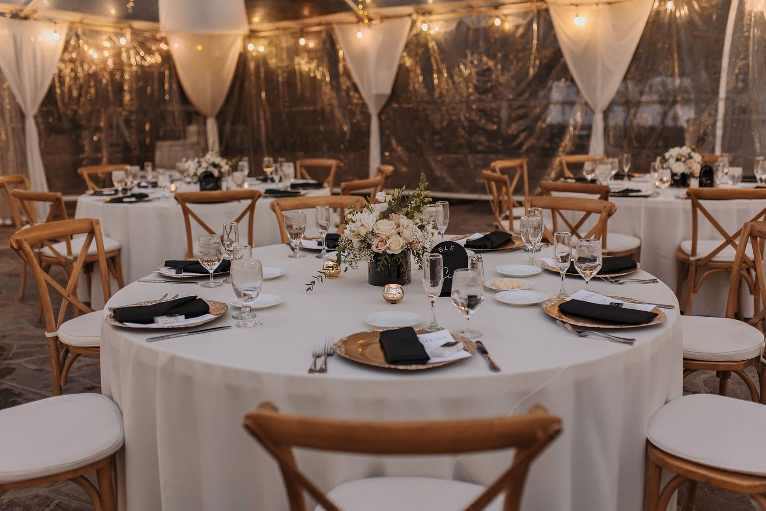 Elegant wedding reception venue with round tables, crossback chairs, and romantic string lights overhead.