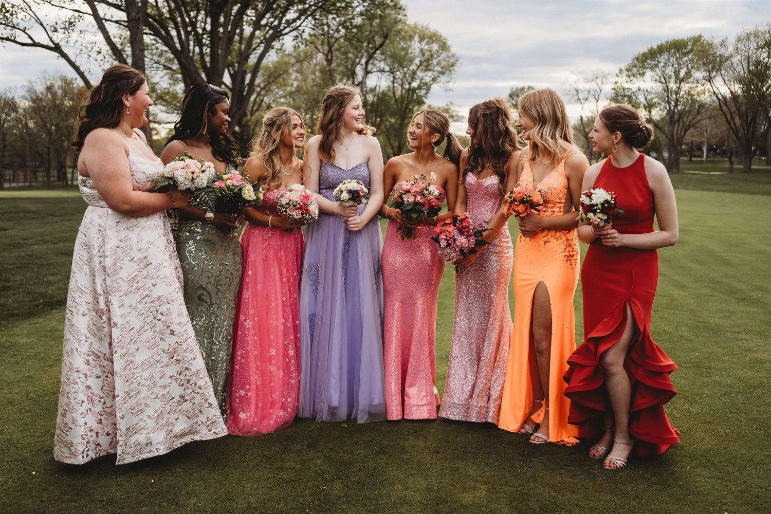 A group in colorful dresses stands together outdoors on a grassy field during a special event. prom