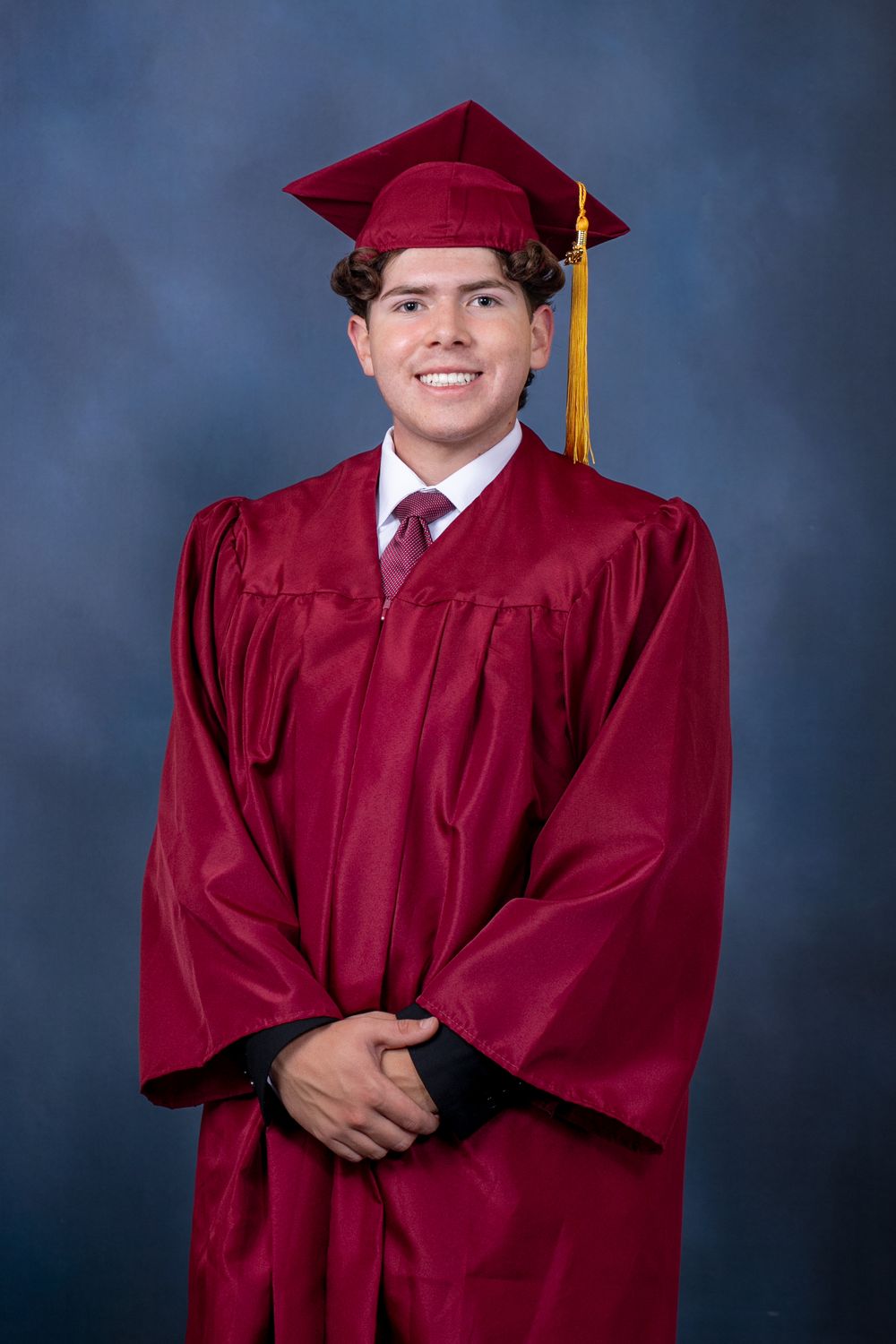 Graduate wearing maroon cap and gown poses for professional portraits against blue backdrop.