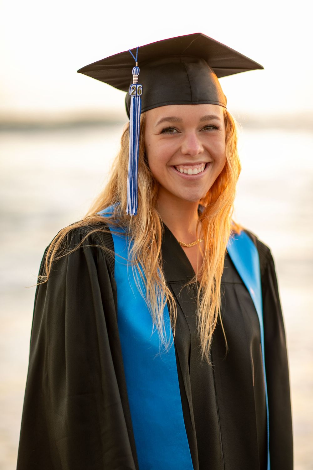 A graduate in traditional black cap and gown with blue stole smiles during sunset graduation photos.