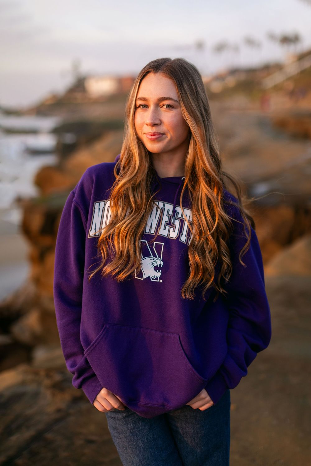Someone wearing a purple Northwestern University sweatshirt poses on rocky coastal terrain at sunset.
