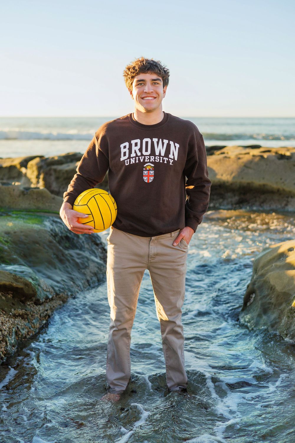 Person wearing Brown University sweatshirt stands on rocky coastal shoreline holding yellow water polo ball at sunset.
