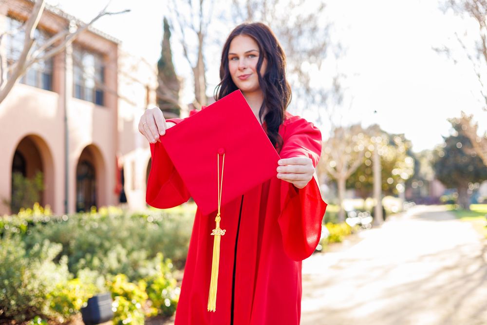 A person in a vibrant red graduation gown walks through a sunny campus courtyard with stone buildings in the background.