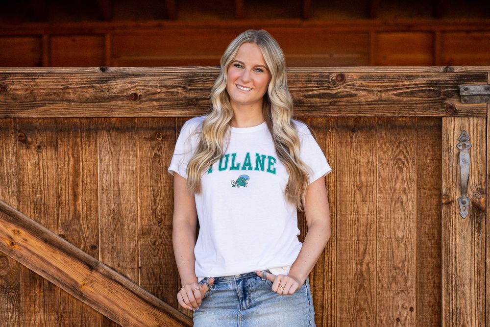 Someone wearing a white Tulane t-shirt and jeans poses against a rustic wooden wall.