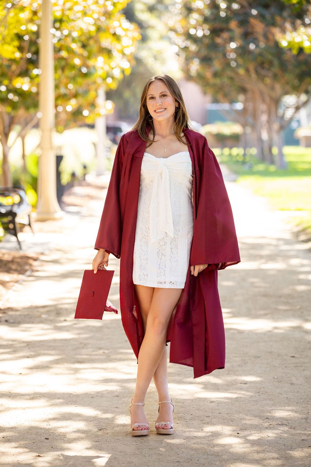 A figure in a burgundy graduation gown and white lace dress poses on a sunlit pathway surrounded by trees.