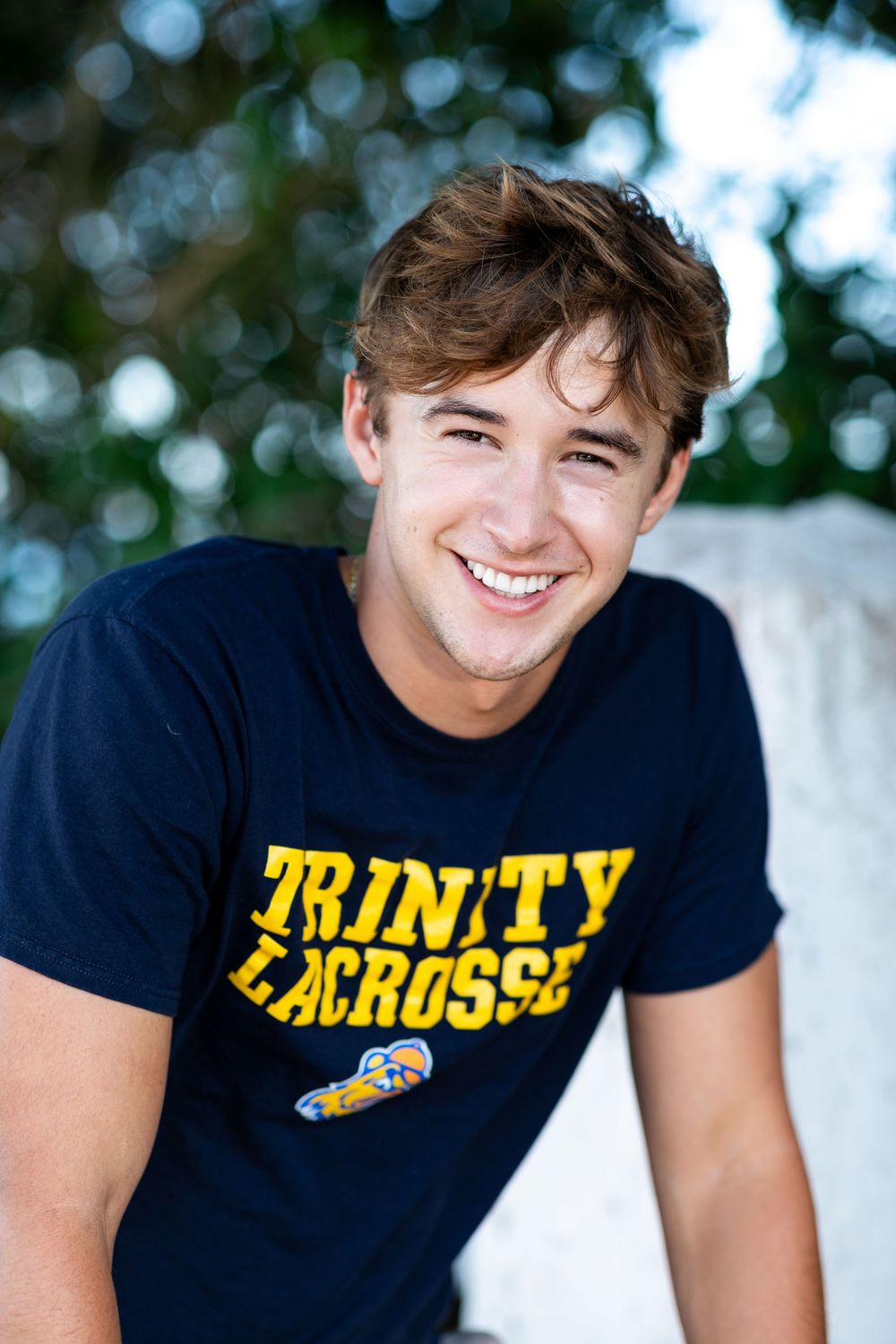 A student athlete wearing a navy Trinity Lacrosse t-shirt smiles outdoors against a blurred natural background.