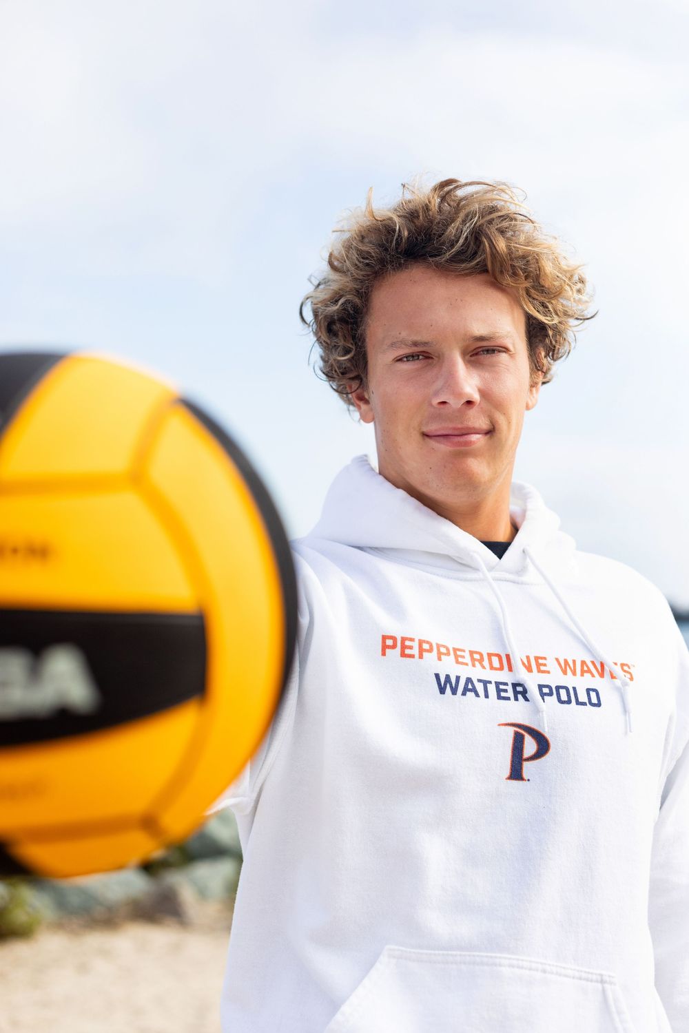 A water polo player in a white Pepperdine Water Polo hoodie stands with a yellow ball on a sunny day.
