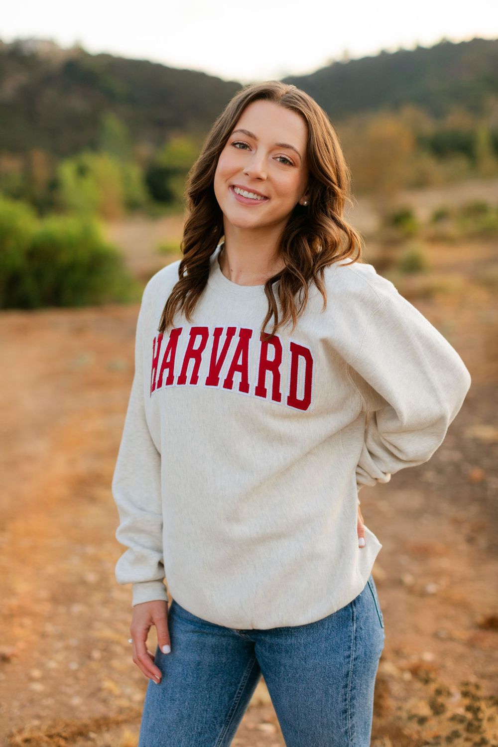 A Harvard sweatshirt worn with blue jeans in an outdoor setting with hills and greenery in the background.