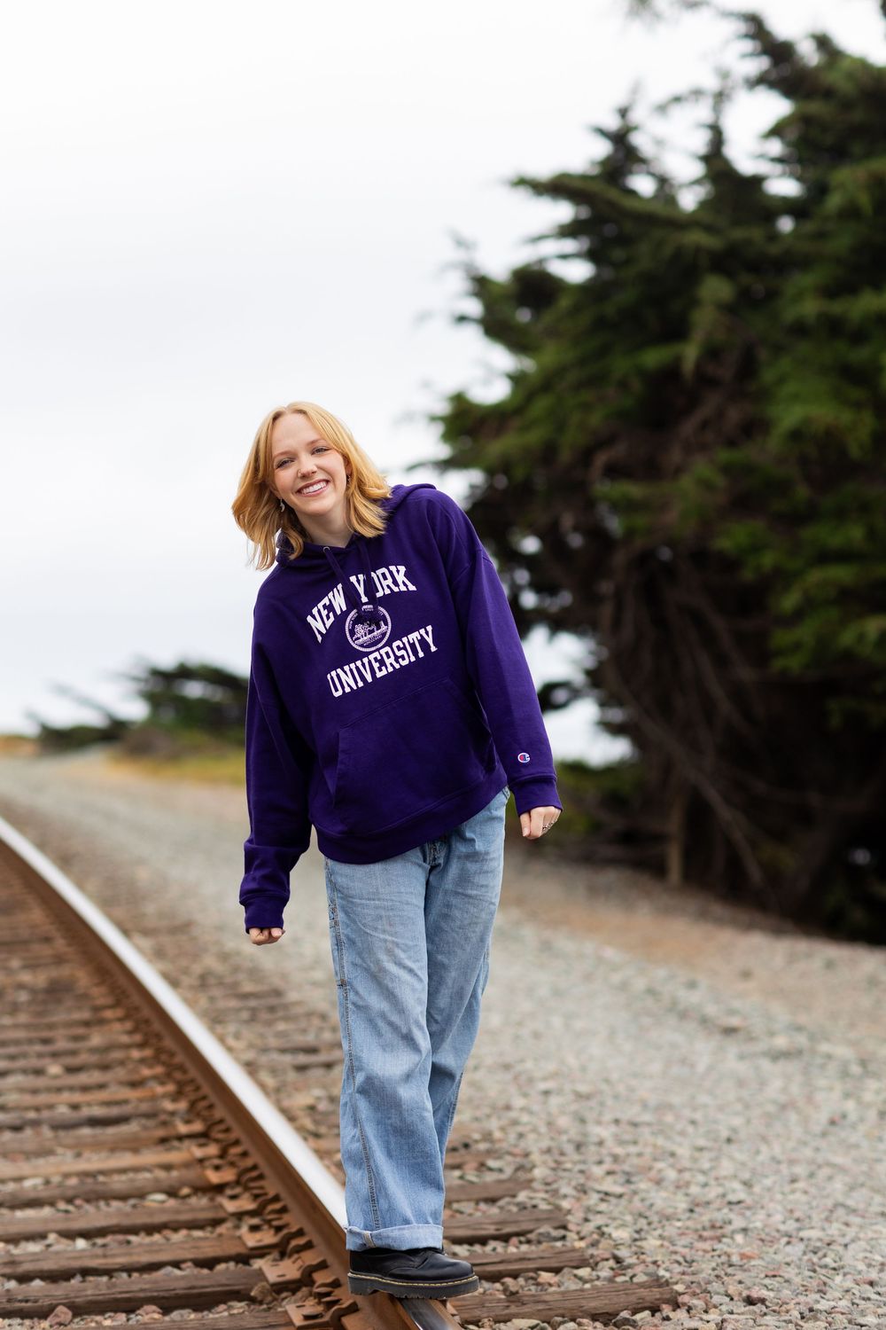 A person in a purple sweatshirt and jeans stands playfully on train tracks surrounded by trees.