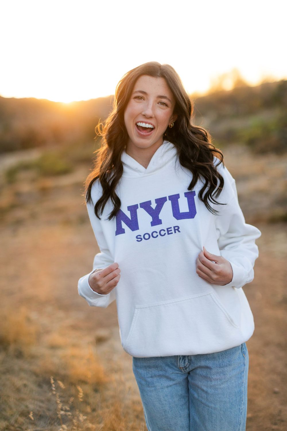 Person wearing white NYU Soccer hoodie and jeans poses outdoors during golden hour sunset.
