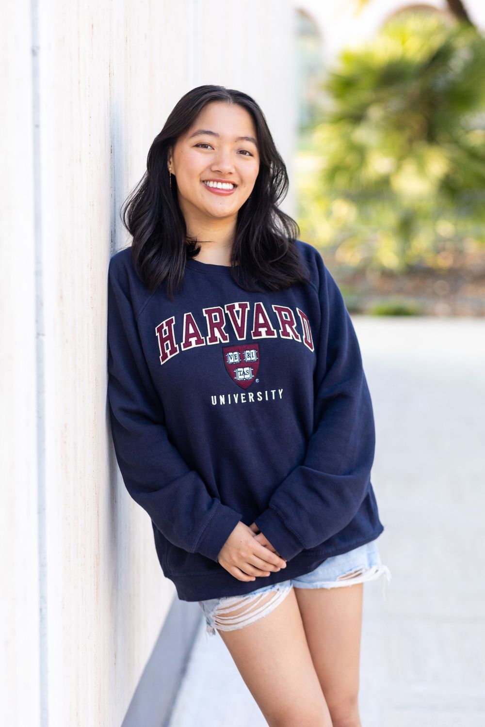 Smiling young woman wearing a navy Harvard University sweatshirt and denim shorts, leaning against a white wall outdoors.