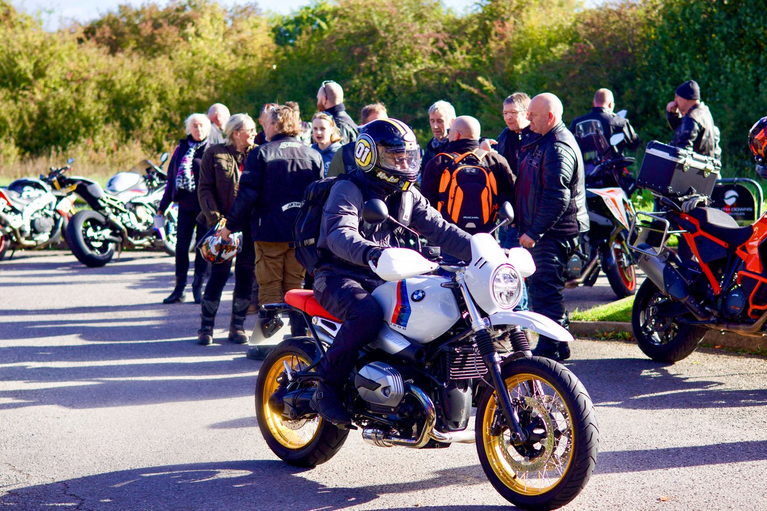 Group of motorcycles parked in a row at a sunny outdoor meetup with riders gathered around them.
