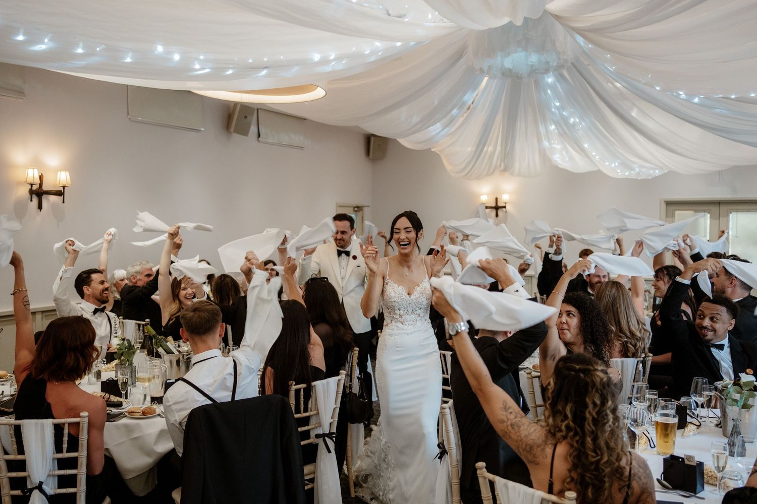 Wedding reception celebration with guests raising glasses for a toast under white draped ceiling and string lights.