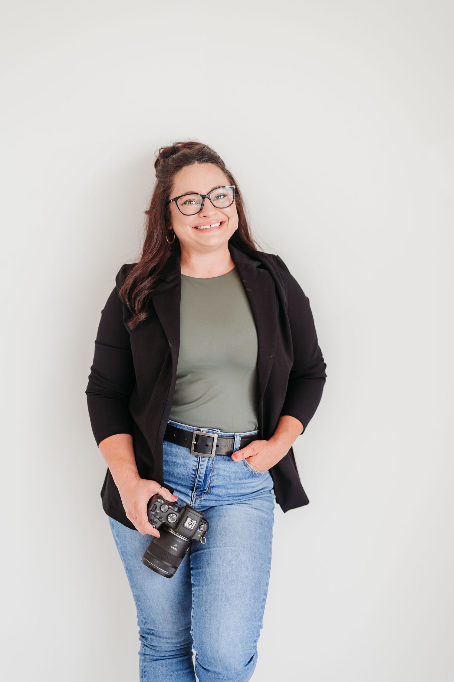 Professional photographer wearing glasses and a black blazer holds a camera against white background.