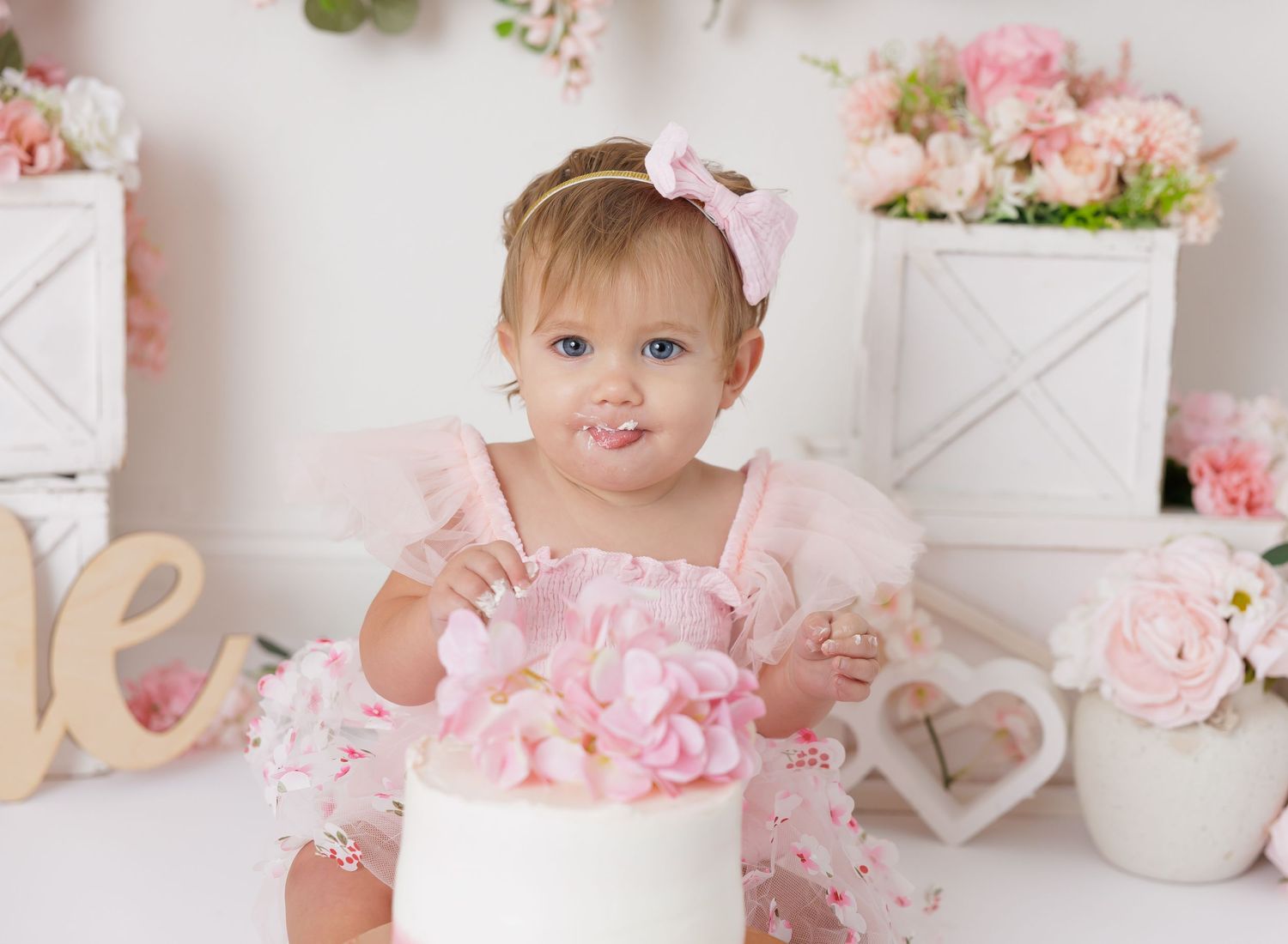 Baby sits in white vintage crib surrounded by rustic farmhouse decor and decorative plants.