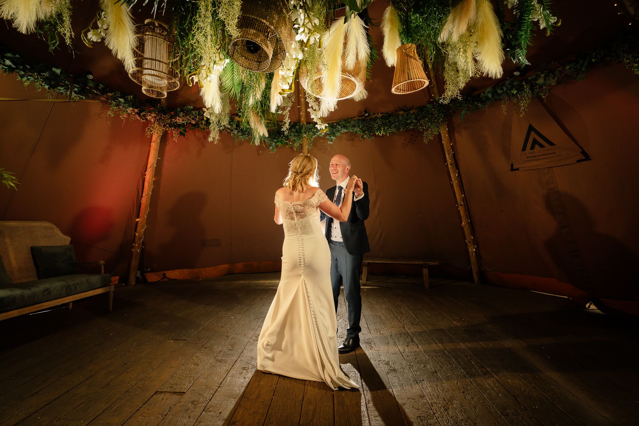 Newlyweds dance under decorative lighting in a rustic wooden reception venue. Woodhill Hall.