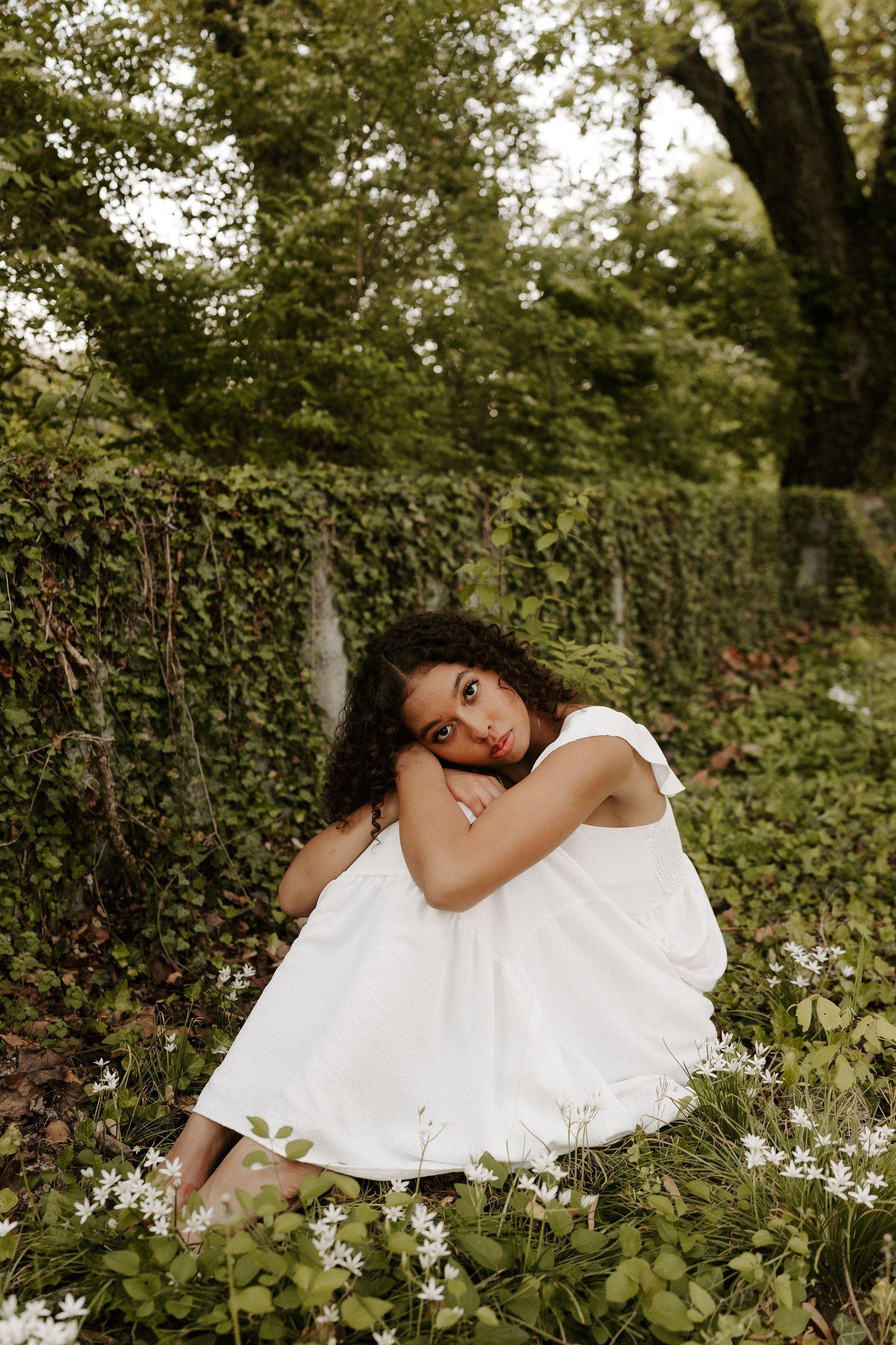 Bride in a white dress sits among wildflowers in a lush green garden setting.
