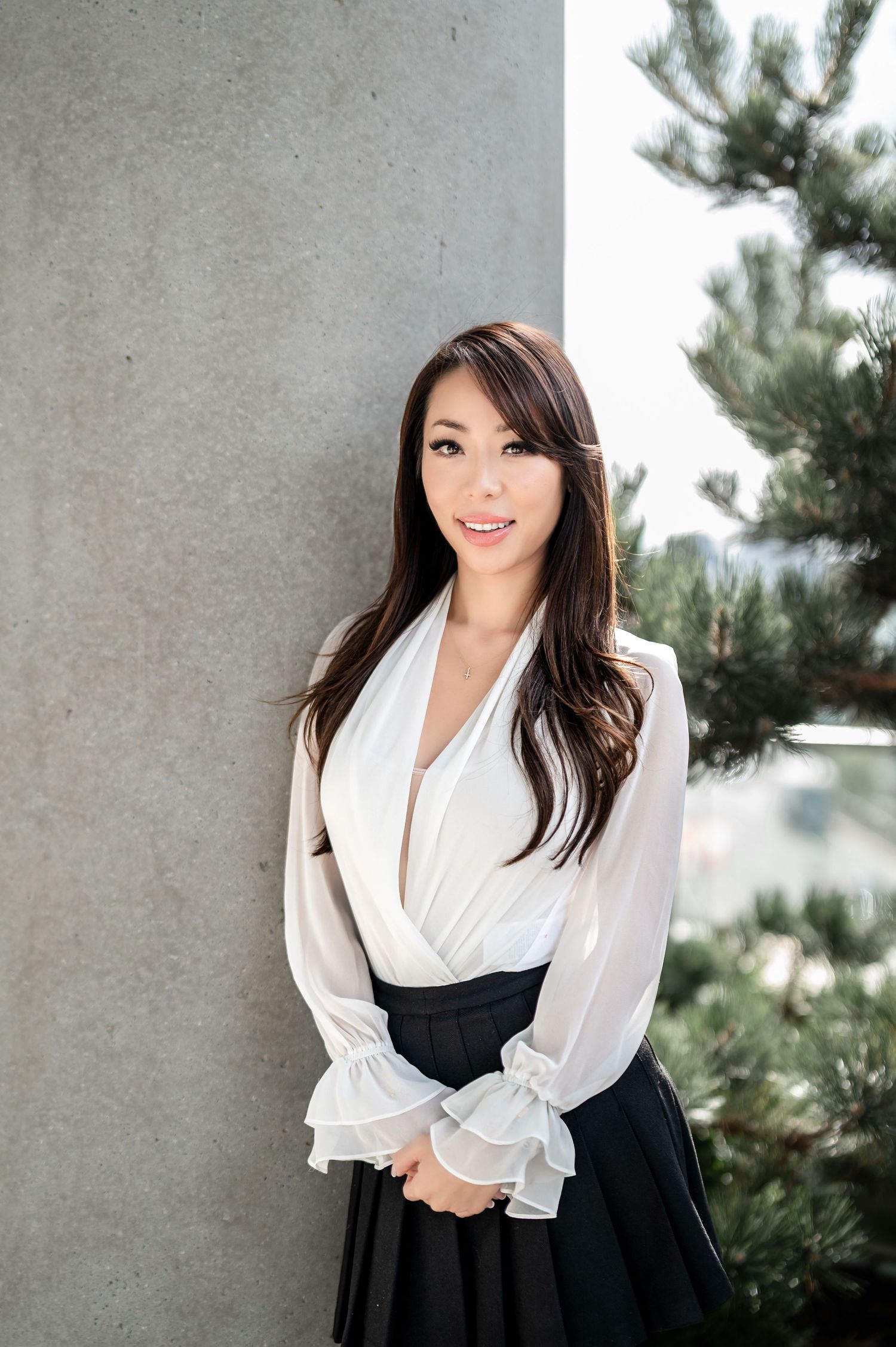 Professional portrait of someone in a white wrap blouse with ruffled sleeves and black skirt standing against a concrete wall.
