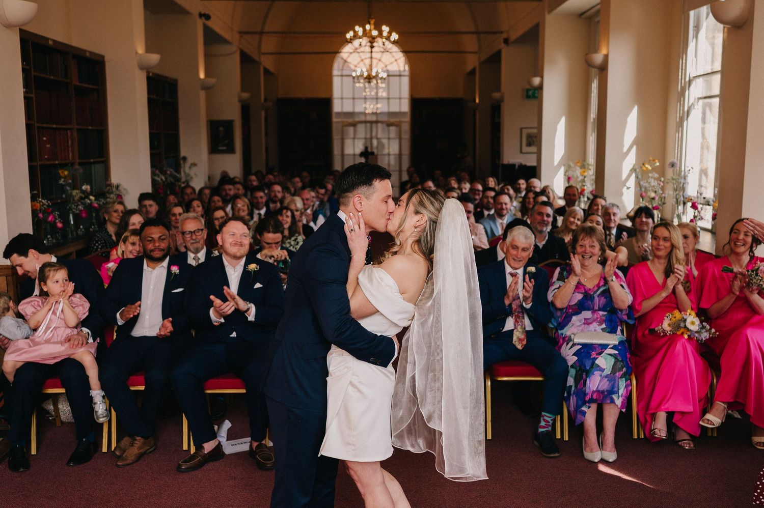 A wedding ceremony in a grand historic hall filled with guests situated in Oxford, England.