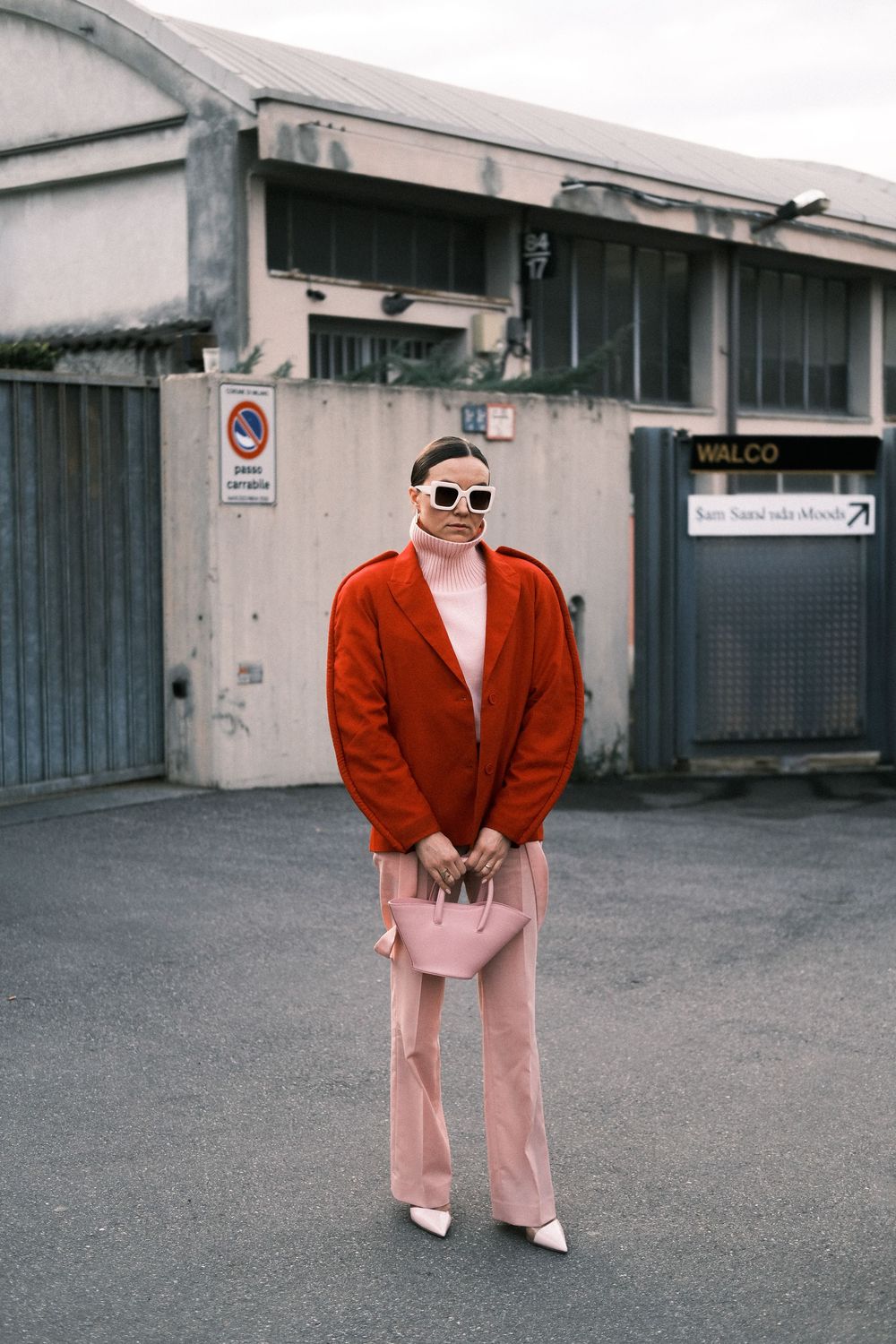 Street fashion look featuring red cardigan, pink trousers, white heels and sunglasses against industrial backdrop.