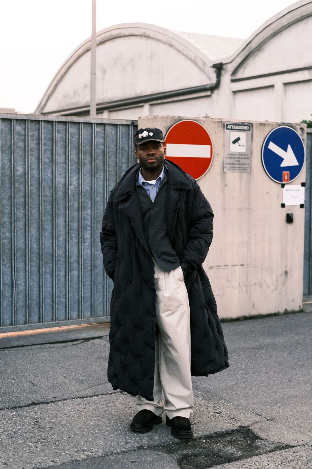 Person in black coat stands against industrial background with curved architectural elements and geometric shapes.