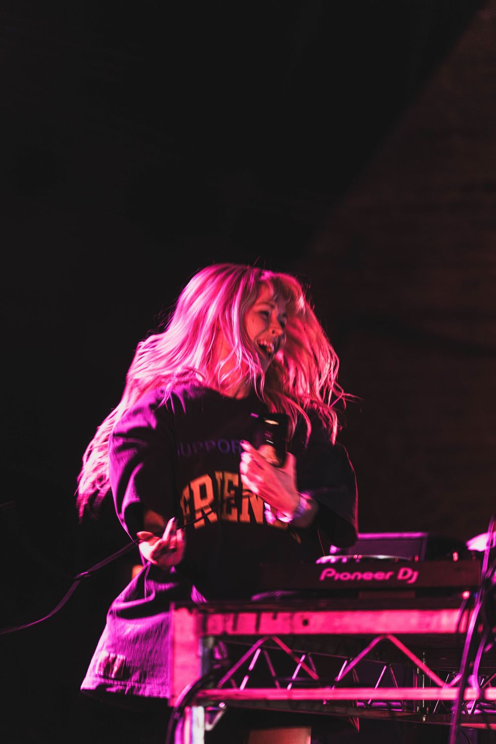 A silhouetted performer illuminated by vibrant pink stage lighting leans over electronic music equipment during a show.
