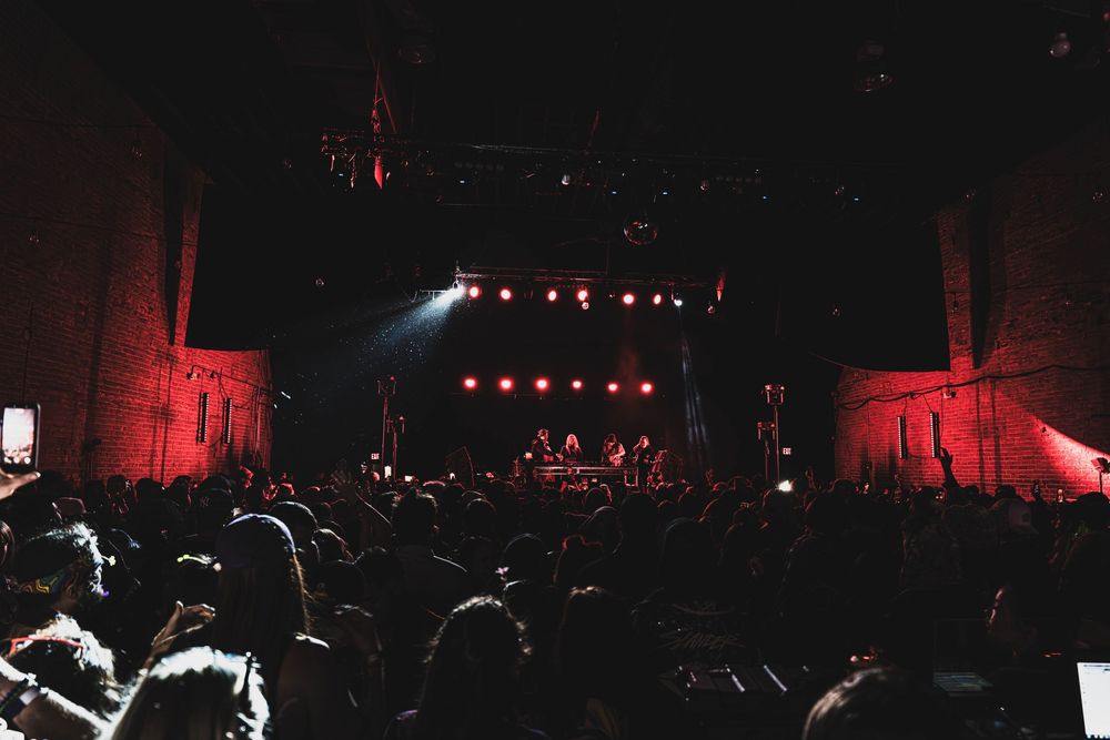 Silhouetted concert crowd watches performers on stage with dramatic red lighting and spotlights in dark venue.