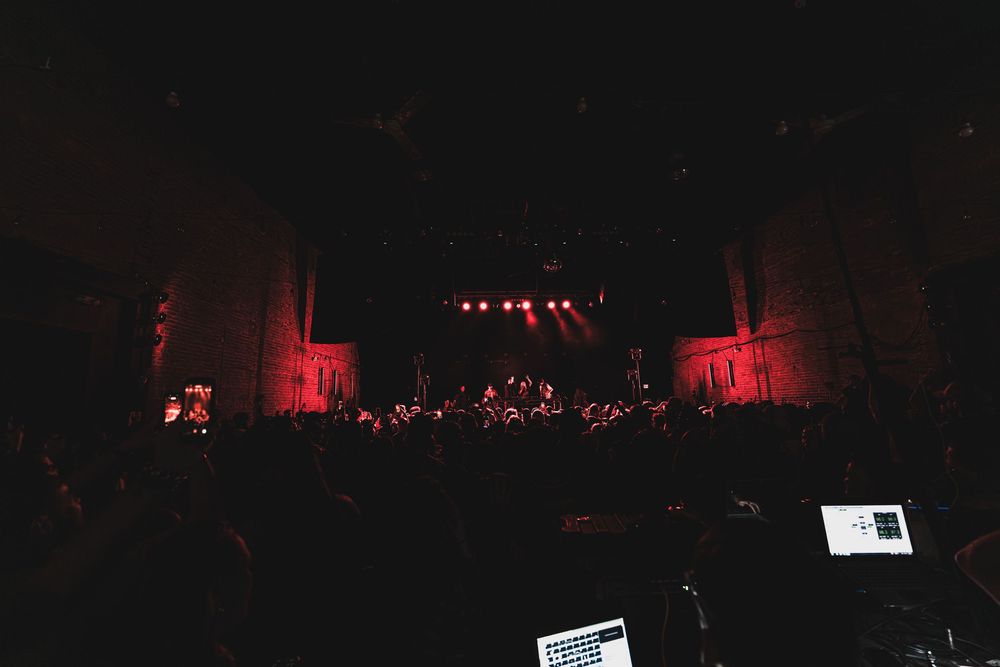 A dark concert stage illuminated with dramatic red lighting and silhouetted figures performing on stage.