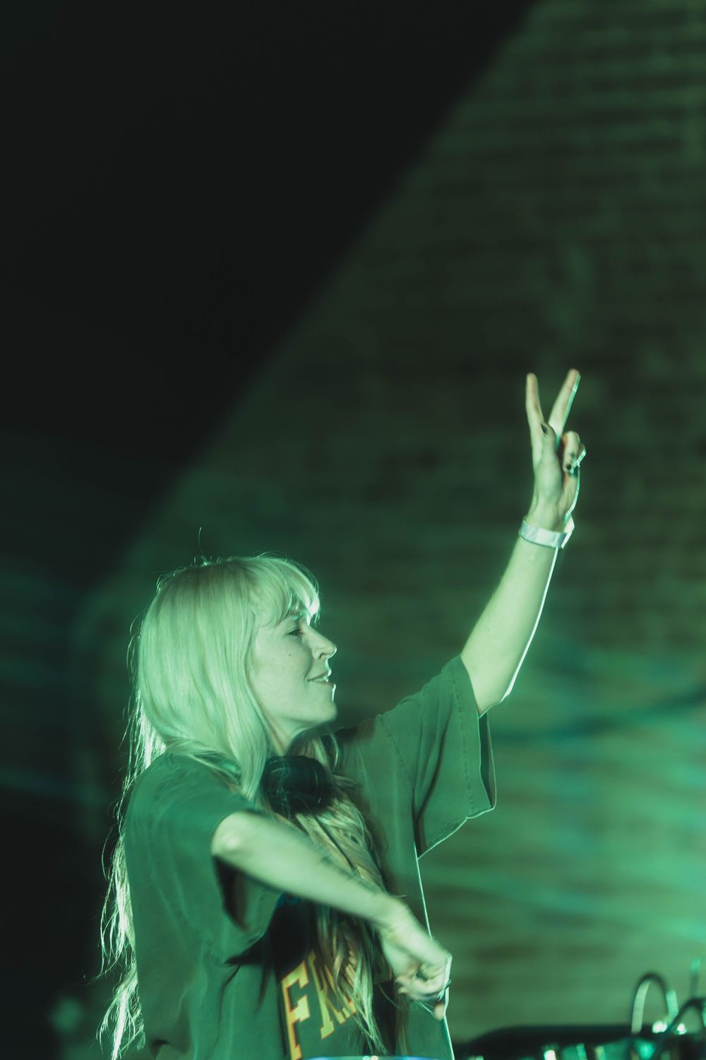 DJ performs at nightclub with raised hand making peace sign while bathed in green lighting during performance.