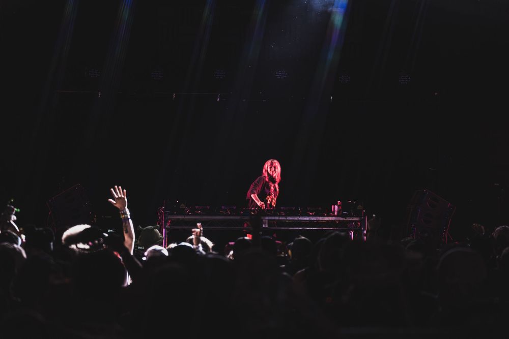 Concert performer plays piano on dark stage with raised hands of audience members visible in silhouette.