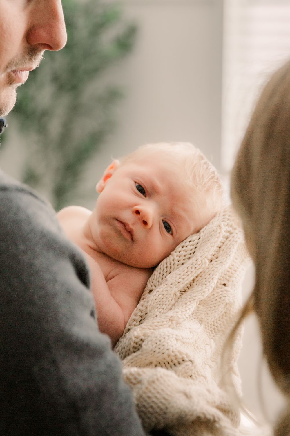 A baby wrapped in a cream knit blanket gazes upward while being held in a tender moment.