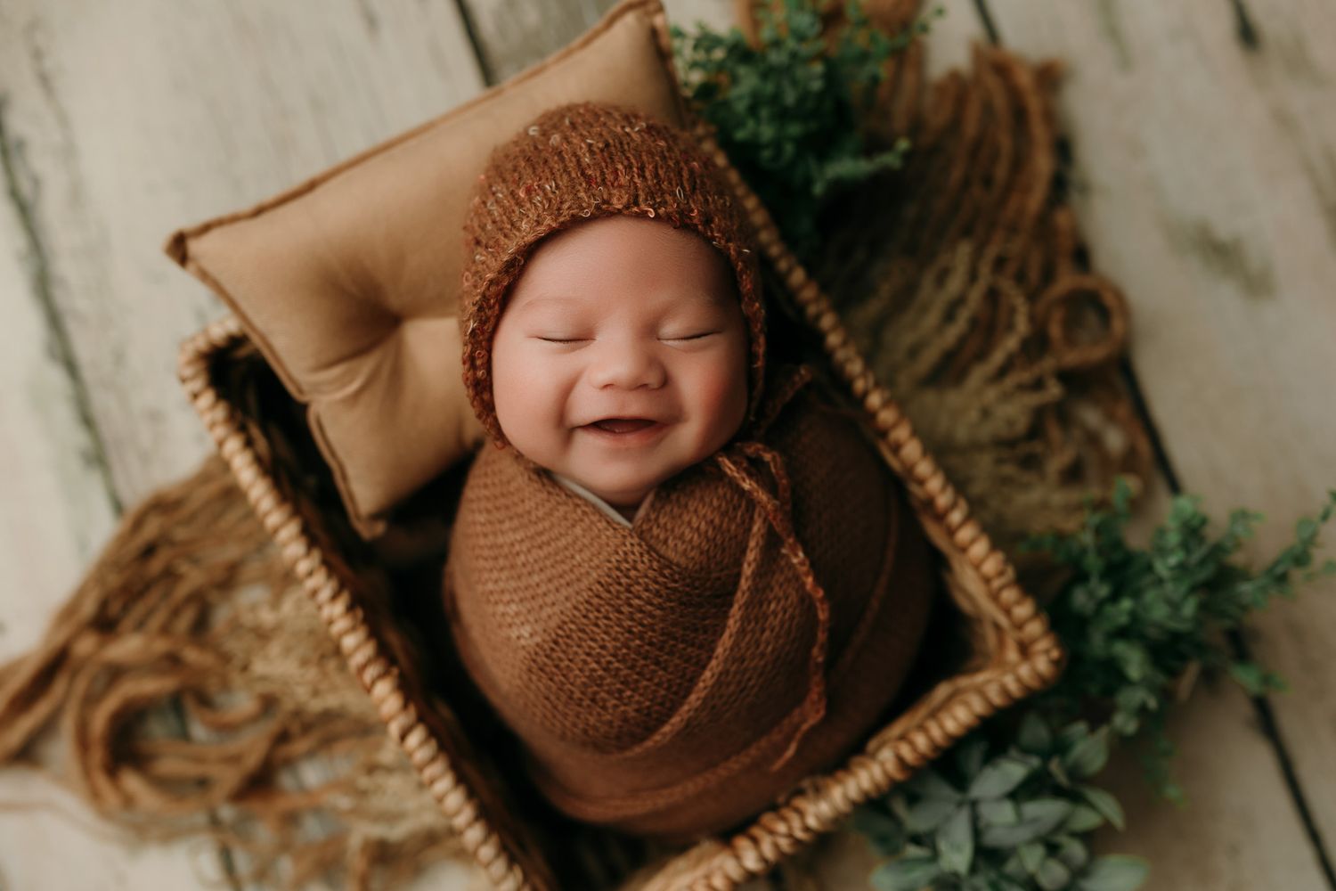 Newborn wrapped in brown knit swaddle and matching hat smiles while lying in woven basket with greenery.