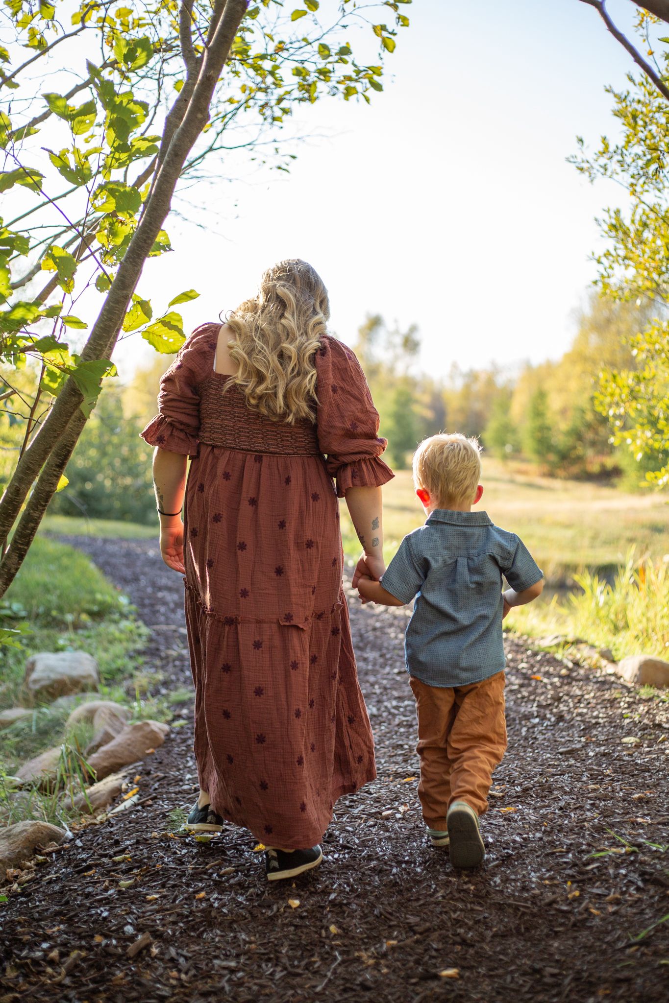 Parent and small child walk together along a wooded path in autumn sunlight, holding hands and exploring nature.