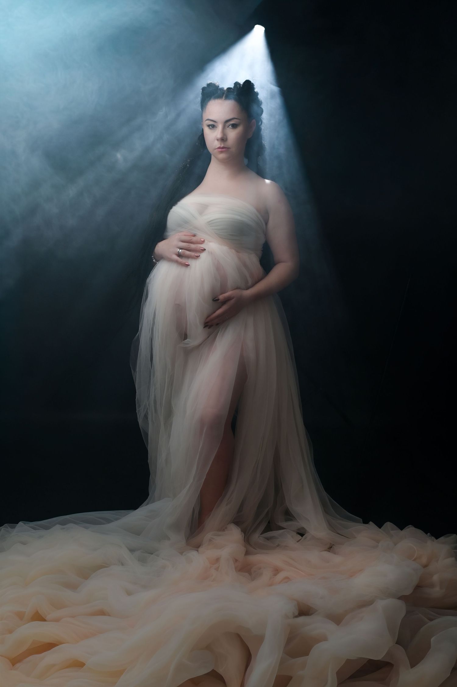 Ethereal maternity photo featuring flowing tulle gown and dramatic lighting with smoke effects in dark studio setting.