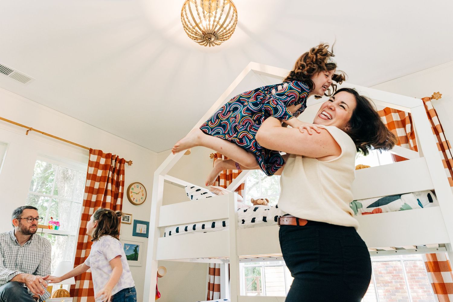 A mom and daughter dances together in a bright, sunlit room with white walls and orange curtains in the background.
