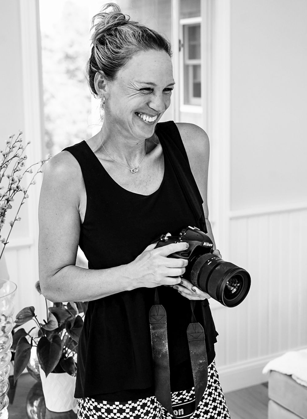 Smiling female photographer holding a Nikon camera indoors in a candid black and white portrait.