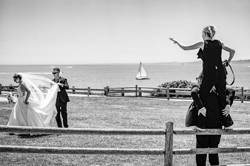 Wedding photographer standing on shoulders to direct bride and groom near ocean fence in black and white.