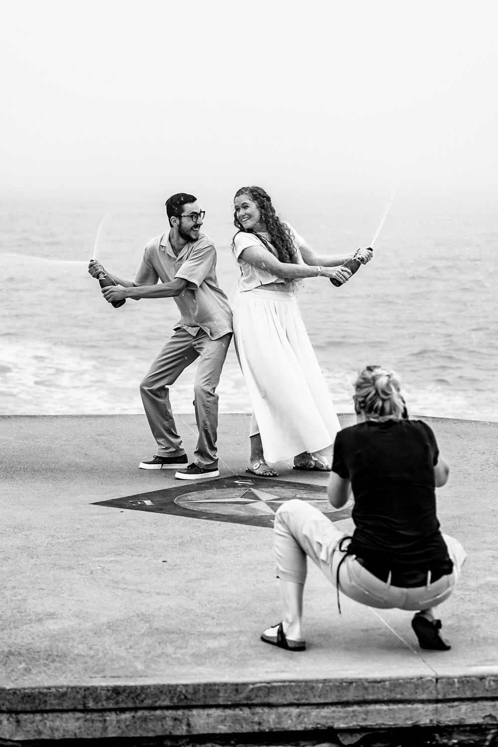 Photographer crouching to capture couple dancing with sparklers by the ocean in black and white.